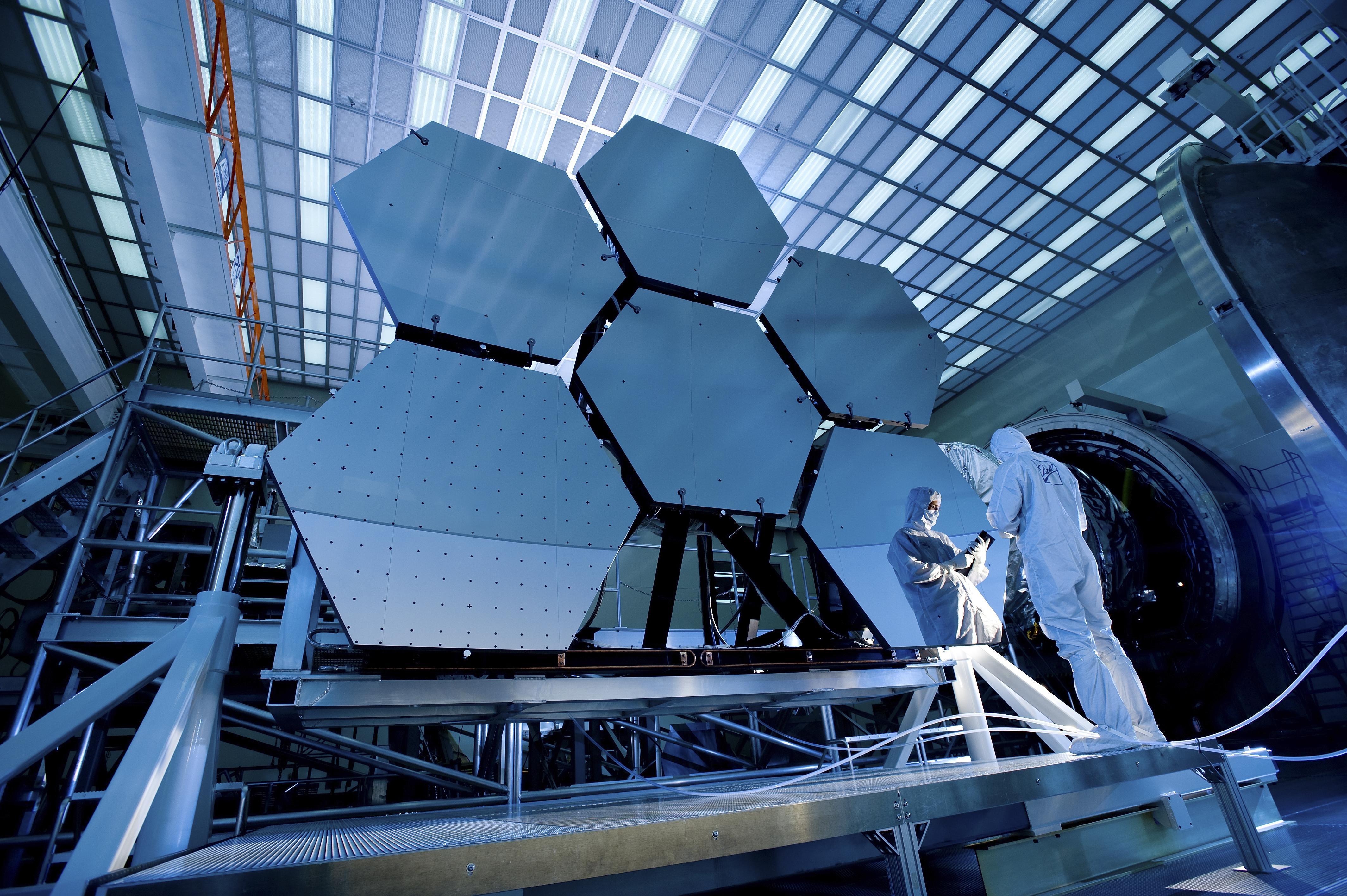Hexagonal mirror segments of a large space telescope under assembly in a cleanroom, with technicians in white suits on a raised platform.